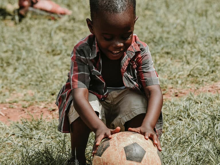 A young child in a plaid shirt and shorts is joyfully playing with a soccer ball on a grassy field. The child is smiling and crouched down, holding the ball with both hands.
