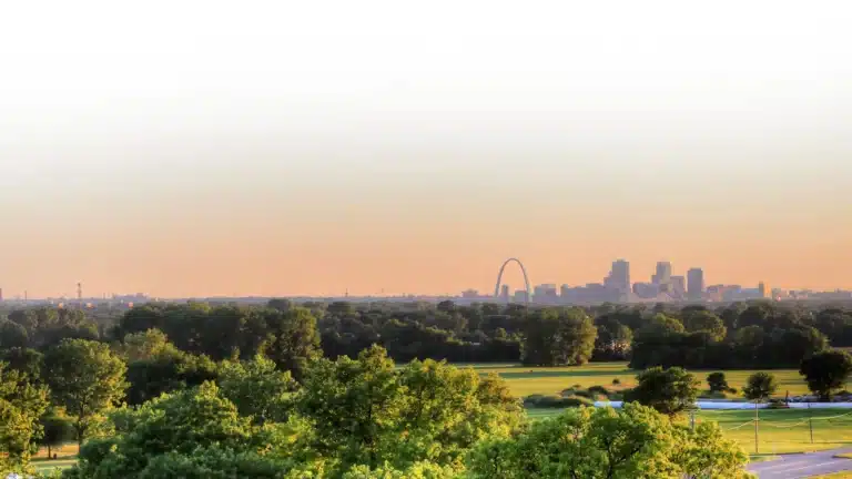A panoramic view of a city skyline during sunset highlights the prominent arch and clustered high-rise buildings. In the foreground, lush green trees and open fields fill the landscape under a clear sky.
