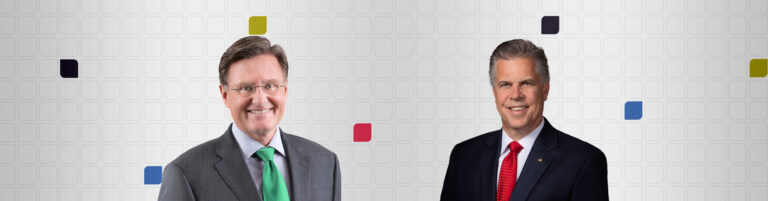 Two men in suits are smiling against a geometric background with small colored squares. The man on the left wears glasses and a green tie; the man on the right sports a dark suit with a red tie.