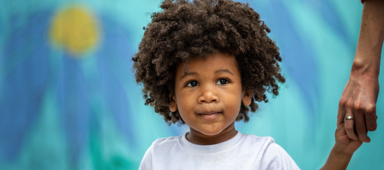 A child with curly hair in a white shirt holds an adult's hand against a vibrant, painted background, showcasing a large blue flower with a yellow center.