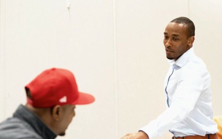 Two men are interacting indoors. One man, wearing a white shirt, is standing and gesturing, while the other man, in a red cap, is seated. They appear engaged in conversation against a plain white background.
