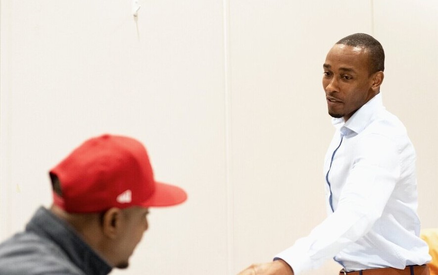 Two men are interacting indoors. One man, wearing a white shirt, is standing and gesturing, while the other man, in a red cap, is seated. They appear engaged in conversation against a plain white background.
