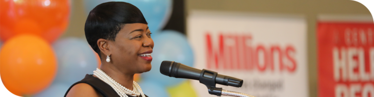 Michelle Tucker, with short black hair and pearl jewelry, smiles while speaking into a microphone. They are standing in front of colorful balloons and banners with text partially visible.