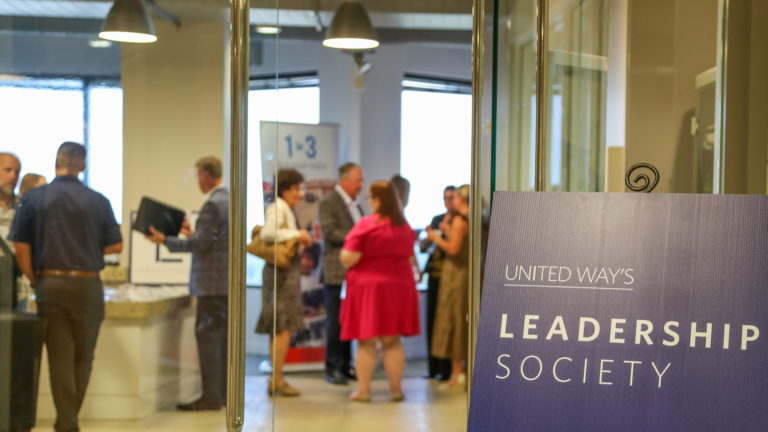 A group of people gather in a modern, well-lit office setting for a United Way event. A prominent sign reads "United Way's Leadership Society." The attendees are actively engaged in conversations, some holding documents.