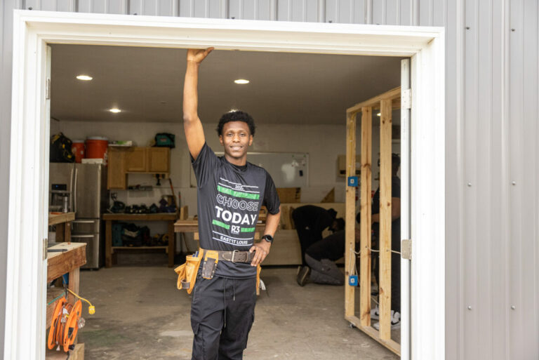 A person wearing a tool belt stands in the entrance of a workshop in St. Louis. They are smiling, with one arm resting on the doorway. Inside, another person is working amidst construction materials and unfinished walls.