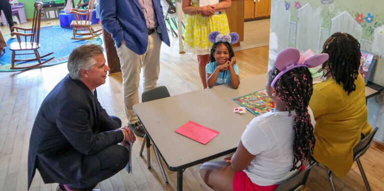 A man in a suit kneels beside a table of smiling children wearing pink and purple mouse ears. Two women stand nearby, engaging with the group in this community-focused setting. The colorful room is filled with playful decorations and a wooden floor.
