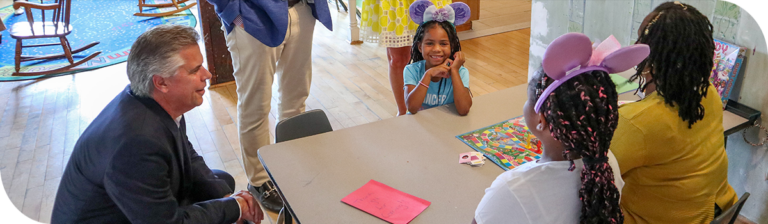 A man in a suit kneels beside a table where three children wearing colorful clothing and headbands are seated, smiling and playing a board game with an adult. This lively scene is set in a bright room with a colorful rug and open space in the background.