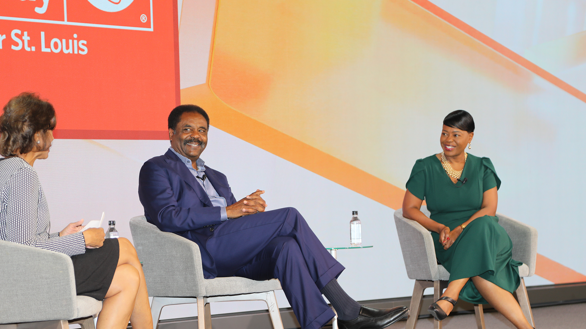 Three individuals are seated on stage engaged in a community panel discussion. The two on the right are smiling, seated in gray chairs with small tables that hold water bottles. A large screen behind them displays part of a logo and text.
