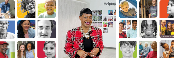Michelle Tucker, in a red and black patterned blazer, stands smiling in a bright room. Surrounding her are photos of diverse people, including children and adults, displayed on the walls.