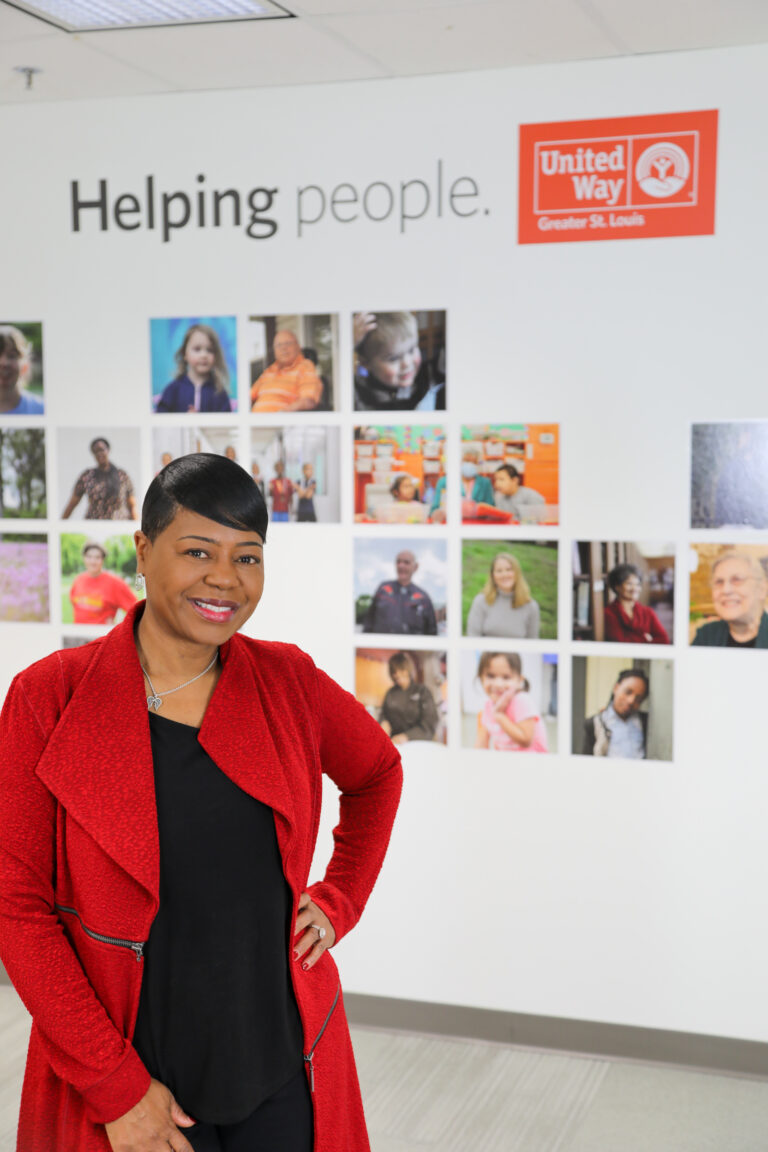 Michelle Tucker, in a red jacket, stands smiling in an office with a wall display behind her. The display features photos of various people and the text "Helping people," along with the United Way Greater St. Louis logo, highlighting volunteer opportunities with this renowned non-profit organization.