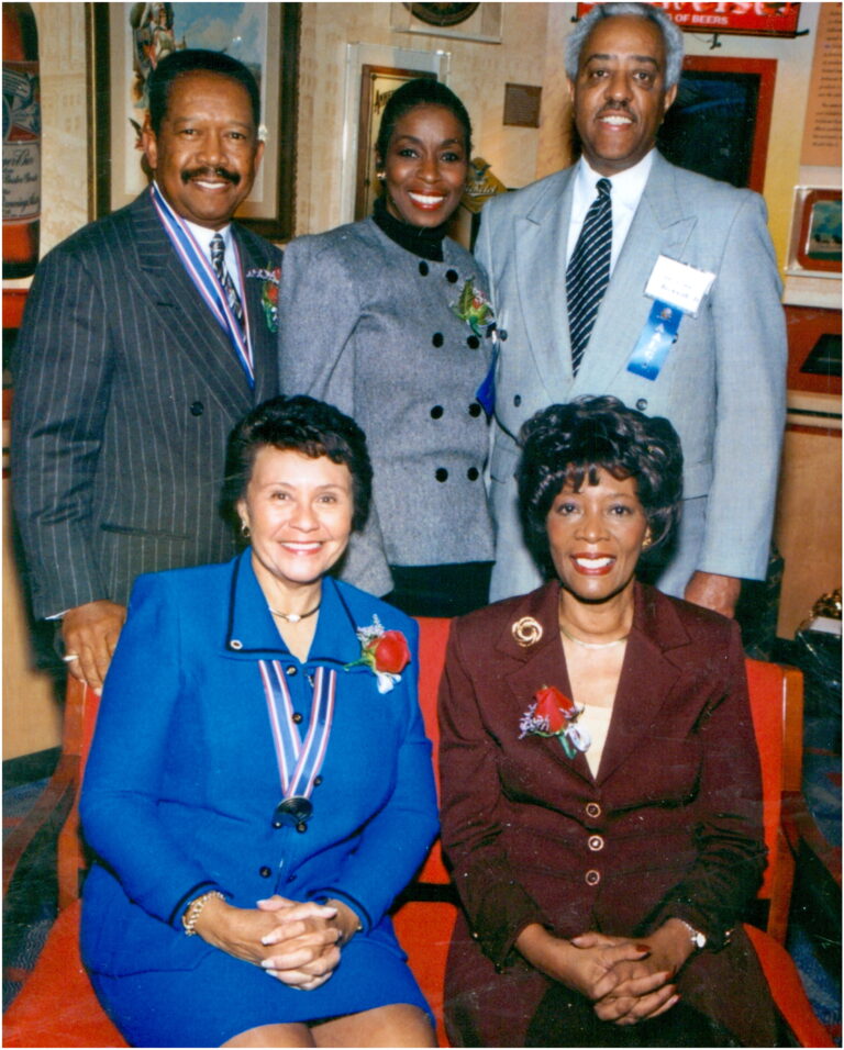 A group of five people poses together indoors, showcasing a sense of community. Two women are seated in front, wearing blue and maroon suits. Three others stand behind them, all in suits and smiling warmly. The background features captivating artwork on the walls.
