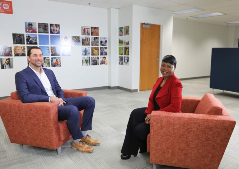 Two people are seated in orange chairs in an office setting, engaged in conversation about community initiatives. The wall behind them is adorned with a collage of colorful photos featuring various people and outdoor scenes. A wooden door is visible to the right, complementing the warm atmosphere.