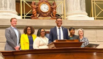 A group of six people, embodying a sense of community, stands together behind a podium in a formal setting. There is a large clock and ornate decorations in the background. The individuals are smiling and dressed in business attire, reflecting camaraderie and professionalism.