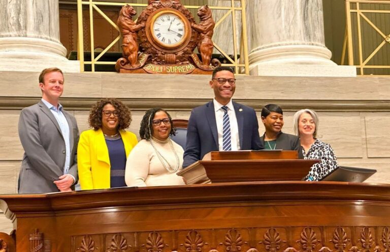 A group of five people stand behind a podium inside a large hall, adorned with ornate decorations and a notable clock behind them. They are smiling and dressed in business attire.