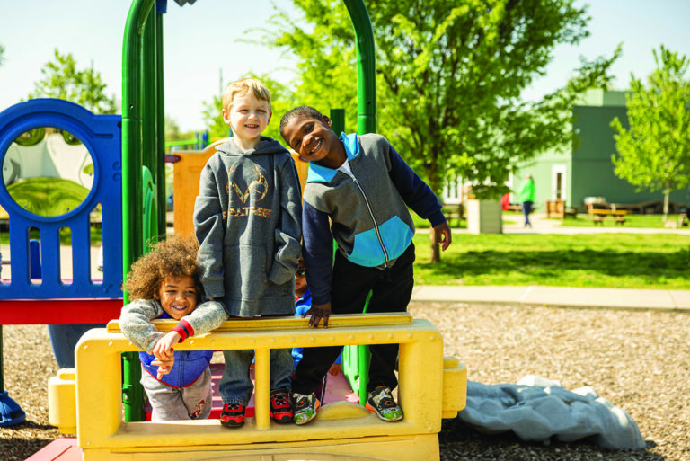 Three children are happily playing on colorful playground equipment outside. The backdrop includes trees and a building, suggesting a sunny day in a community park or playground setting.