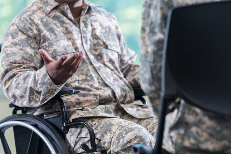 A person in a camouflage military uniform sits in a wheelchair, gesturing as they converse with another individual who is partially visible. The setting appears to be indoors with a blurred background.