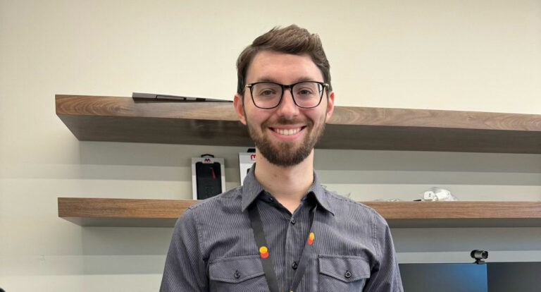 A bearded man wearing glasses and a button-up shirt, stands smiling in a modern and tidy office. Behind him are wooden shelves with various items, including a phone and small objects. The setting reflects the professional yet inviting atmosphere.