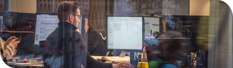 A group of people works at a shared desk with multiple computer monitors. One person is typing, while another is on the phone. The scene is viewed through a window, reflecting light and outdoor elements, suggesting a classroom.