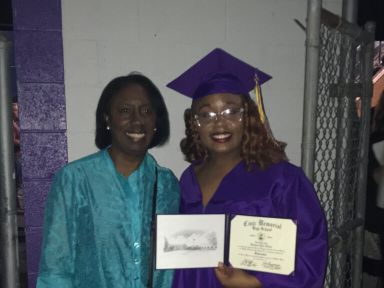 A graduate in a purple cap and gown smiles while holding a diploma, standing beside another person wearing a turquoise outfit. They are indoors, with a white and purple wall in the background, possibly capturing a celebratory moment at an event in St. Louis.