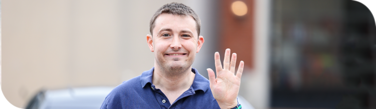 A man wearing a blue polo shirt smiles and waves at the camera. The background is blurred, hinting at an urban setting with a glimpse of a vehicle.