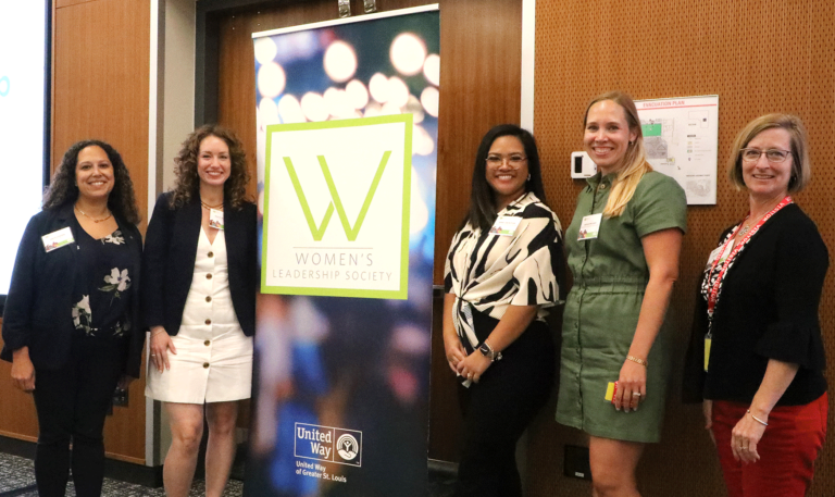 Five women stand smiling beside a "Women's Leadership Society" banner. They are indoors, dressed in business casual attire.