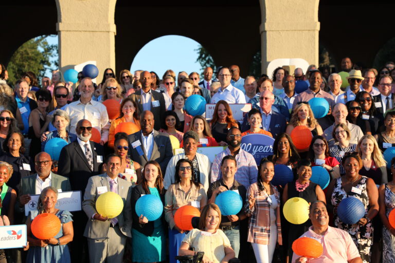 A large, diverse community gathers outdoors, many holding colorful paper lanterns and smiling at an event beneath arches and trees. Some hold signs with writing on them, showcasing a united way of celebrating together.