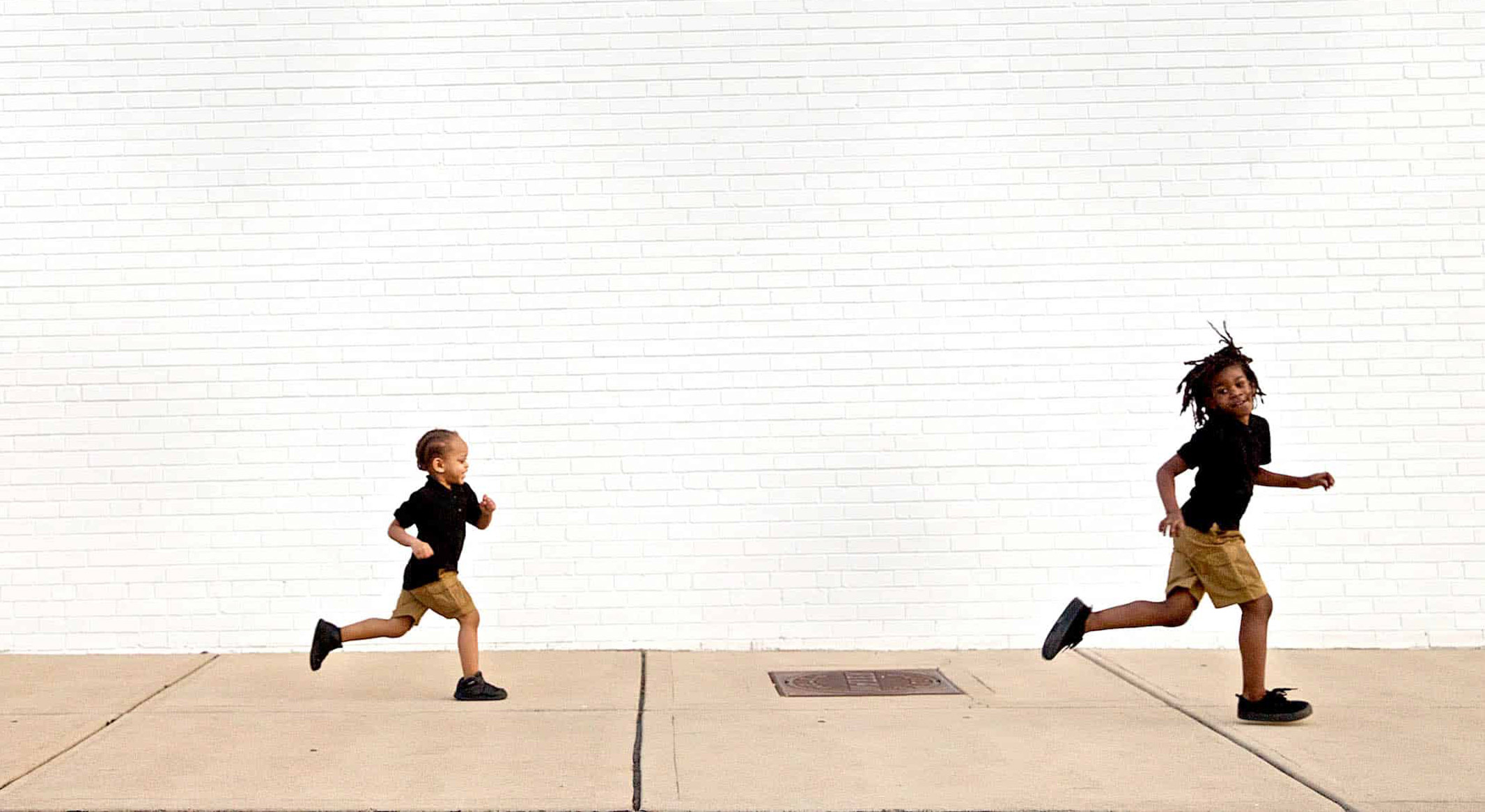 Two kids in black shirts and khaki shorts are running on a sidewalk against a large white brick wall. The older child is ahead, smiling, with dreadlocks flying in the air, while the younger child follows behind with a determined expression.