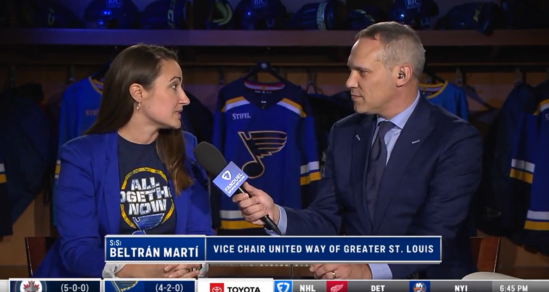 United Way Campaign Vice-Chair, SiSi Beltran Marti, and a man are speaking on stage with a microphone in front of them. Behind them are hockey jerseys. The woman is wearing a blue T-shirt and blazer, and the man is in a suit. Text on the screen displays their names and affiliations.