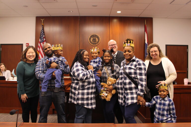 A family of 7 stands in a court room, flanked by two social workers with a judge standing behind them.