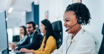A smiling woman sits in a call center with a headset on, looking at a computer screen. Others are seen doing the same in the background.